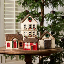 A set of miniature Christmas village houses and barns displayed on a round wooden pedestal in front of a decorated Christmas tree and window shutters.