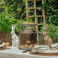 Decorative table setting with Easter-themed items including rabbit figurines, eggs, two-tier metal stand and a wooden ladder in a garden.