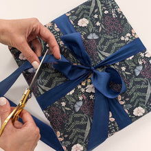 Person cutting a blue ribbon on a floral-patterned gift box with scissors.