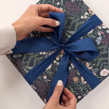 Gift box with floral wrapping paper and a blue ribbon being adjusted by hands.