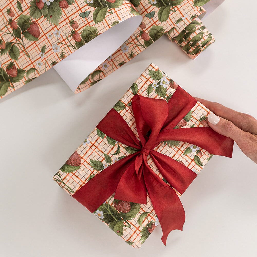Gift box wrapped in strawberry patterned paper with a red ribbon, on a white surface.
