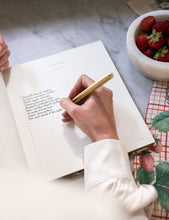 Person writing in a recipe book with a pen, next to a bowl of strawberries on a marble surface.