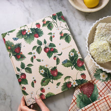 Floral-patterned book held by a hand on a marble surface with lemons and a bowl of lamingtons in the background.