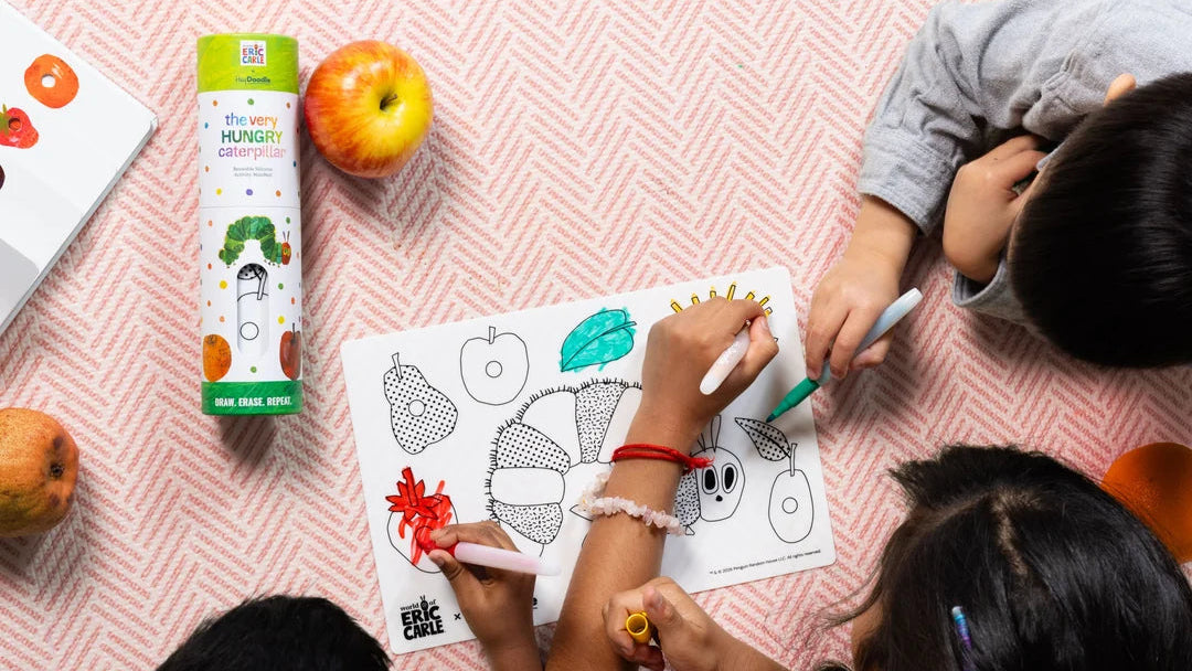 Three children are seen from above drawing on a coloring page with markers while sitting on a pink patterned blanket. A Very Hungry Caterpillar coloring book and other children's items are visible nearby.