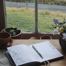 Open notebook with pen on a wooden table near a window with a view of a grassy area.
