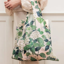 A person in a beige blouse holds a large fabric tote bag with a green and white botanical print featuring hydrangea flowers, set against a white paneled wall.