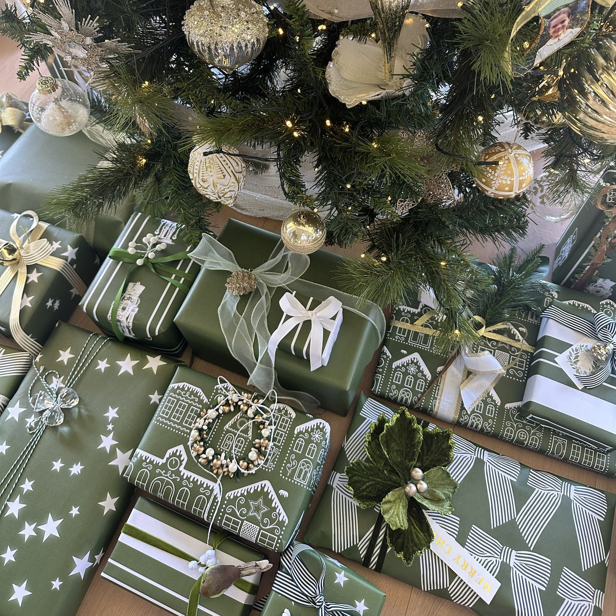 A festive holiday scene showing a decorated Christmas tree with silver and gold ornaments, warm string lights, and wrapped gift boxes underneath. The presents are wrapped in green paper with white patterns, ribbons, bows, and small decorative wreaths.