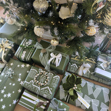 A festive holiday scene showing a decorated Christmas tree with silver and gold ornaments, warm string lights, and wrapped gift boxes underneath. The presents are wrapped in green paper with white patterns, ribbons, bows, and small decorative wreaths.