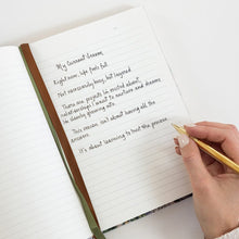 An open lined notebook with handwritten text on the page, a green ribbon bookmark, and a hand holding a gold pen over it on a clean white background.