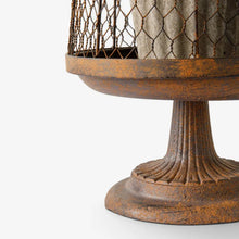 Close-up of a rustic metal pedestal table or stand with a fluted base and wire mesh top, shown on a light gray background.