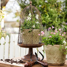A rustic metal birdcage planter displayed on a wooden table in a garden setting, with soil, gardening tools, and pink flowers in the background.
