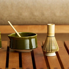 A green ceramic matcha bowl with a wooden spoon and a bamboo whisk resting beside it on a wooden slatted table, with a softly blurred indoor background.