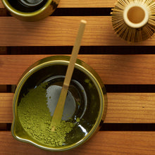 A top-down view of a green ceramic bowl filled with matcha green powder and a bamboo whisk, set on a wooden slatted surface alongside other matching green and gold ceramic tea accessories.
