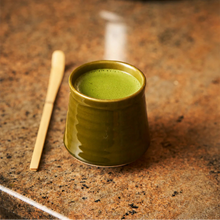 A small glazed ceramic cup filled with bright green matcha tea sits on a speckled brown granite countertop next to a bamboo matcha whisk.