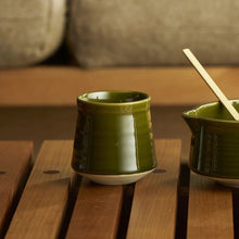 A glossy olive green matcha ceramic cup and matching bowl with a wooden spoon, set on a wooden slatted table with a blurred sofa in the background.