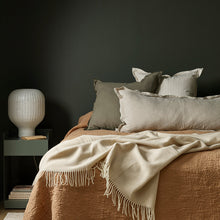 Bed with beige bedding and pillows against a dark wall with a side table and lamp.