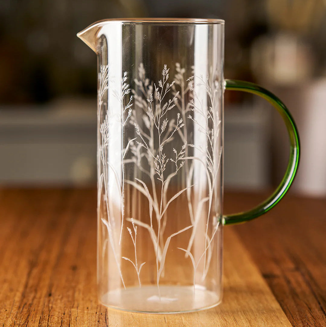 Clear glass pitcher with engraved tree design and green handle on a wooden surface