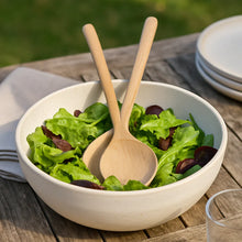 White bowl with green salad and wooden spoons on a wooden table outdoors