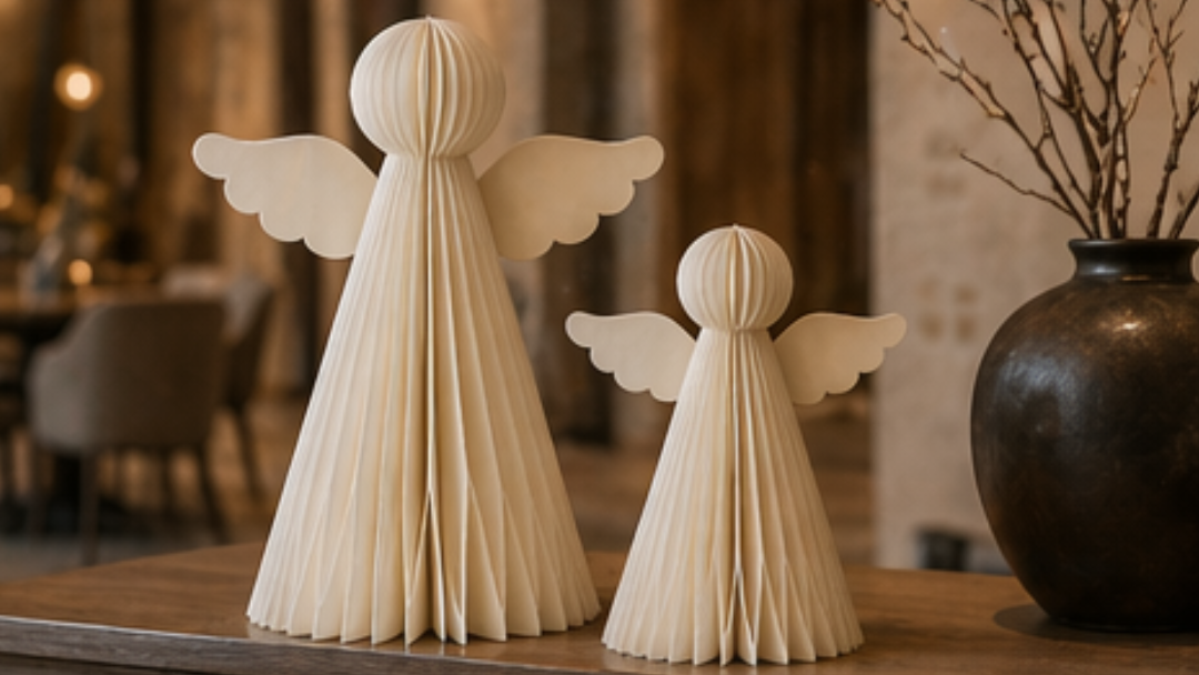 Two cream-colored angel figurines with pleated wings and bodies, displayed on a wooden table in a warm, softly blurred interior setting next to a dark ceramic vase with dried branches.