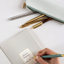 Hand holding a pen with a name card next to a notebook and stationery on a white surface