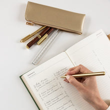 Person writing in a notebook with a gold pen, surrounded by stationery items on a light surface.