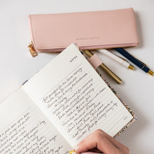 Handwriting in a notebook with a pink pen and pink pencil case on a white surface