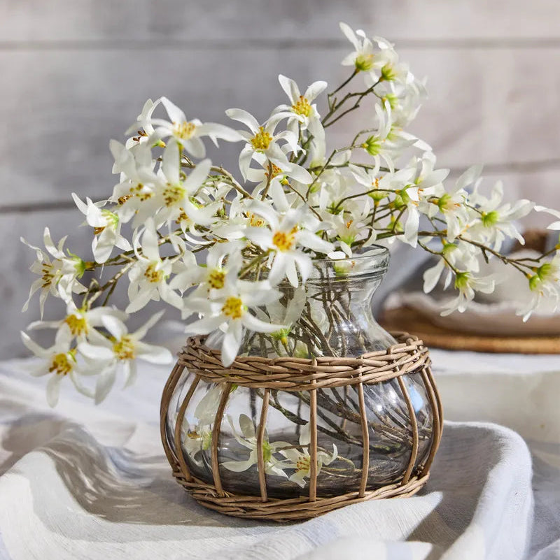 Glass vase with wicker base containing white flowers on a light surface.