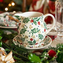A white ceramic Christmas creamer pitcher with a green holly and red berry pattern, resting on a matching saucer on a decorated holiday table with pine branches and warm blurred lights in the background.