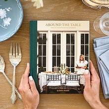 Book titled 'Around the Table' held by hands on a table with floral arrangement and silverware.
