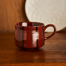 A glossy dark red ceramic coffee mug with vertical black stripes, sitting on a wooden table with a blurred white plate in the background.