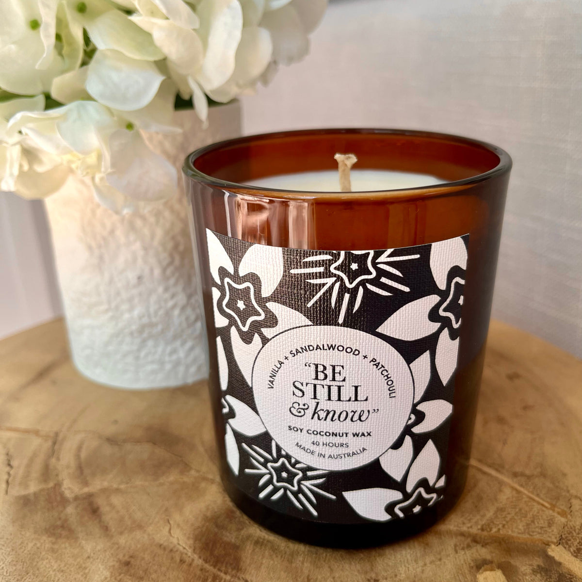 Candle with decorative label on a wooden surface next to a vase of white flowers.