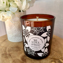Candle with decorative label on a wooden surface next to a vase of white flowers.