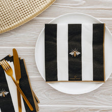 Black and white striped napkin with bee design on a white plate, gold cutlery, and woven mat on wooden surface.