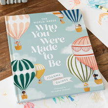 Book titled 'The World Needs Who You Were Made to Be' by Joanna Gaines on a wooden surface with colorful illustrations of hot air balloons.
