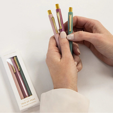 Three pastel-colored retractable pens with gold accents are held between two hands, with their retail box visible on the left against a clean white background.