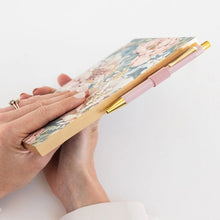 A close-up of a person's hands holding a slim notebook with a pastel floral cover and a pink pen with gold accents resting on top, set against a clean white background.