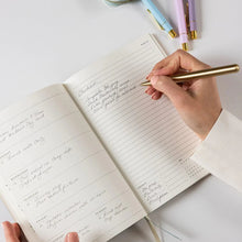 Person writing in a notebook with a gold pen on a light surface.