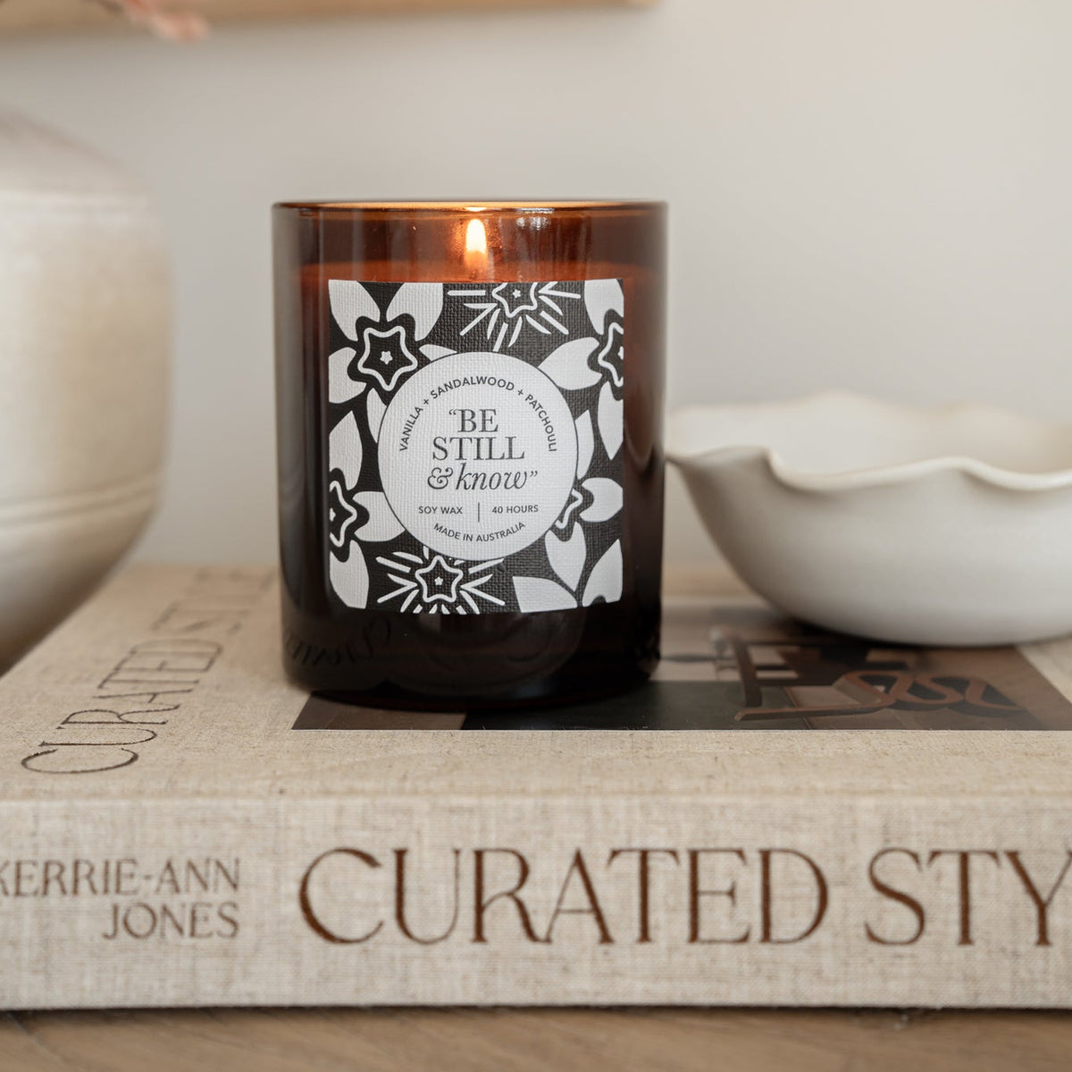 Well Versed Homes Signature Candle with amber glass and label that say 'be still and know', sitting on book with bowl