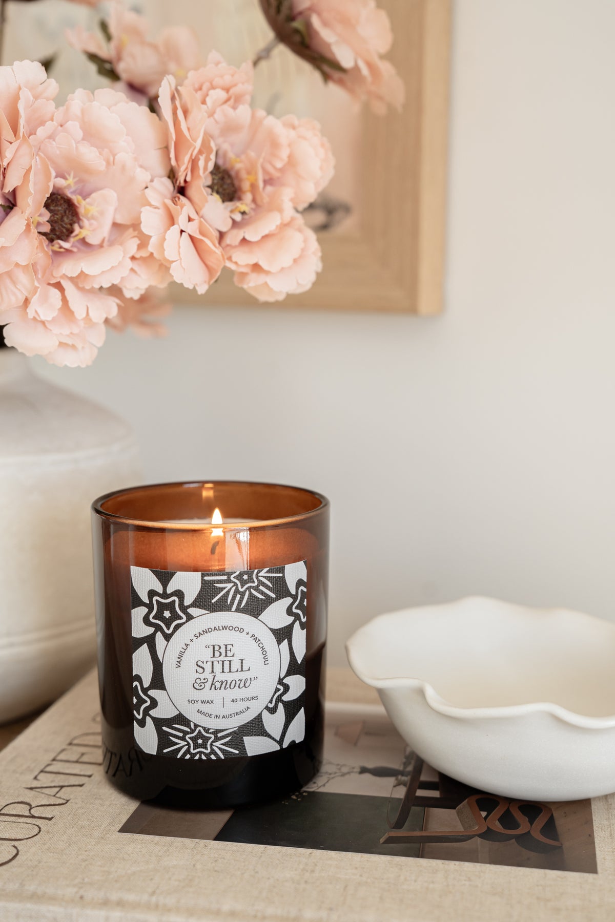 Well Versed Homes Signature Candle with label that says 'be still and know' sitting on book with bowl and flowers