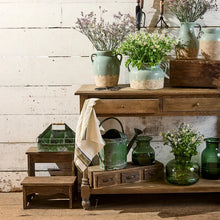 A styled rustic interior scene with a wooden console table holding multiple ceramic vases, green glass containers, a watering can, and dried and white flowers against a white shiplap wall.