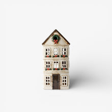 A small decorative house figurine with a white facade, dark brown roof and door, and green holiday wreath details on the roof and windows, shown against a plain white background.