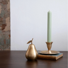 A decorative brass pear-shaped object and a brass candlestick holder with a tall pale green candle, styled on a dark wooden surface against a white wall.