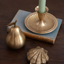 A brass candlestick holder with a light green candle, styled on top of a vintage blue book alongside a brass pear and a brass shell on a dark wooden table.